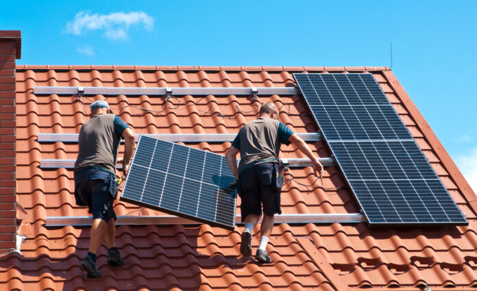 Zwei Arbeiter installieren eine Solarzelle auf einem roten Ziegeldach.