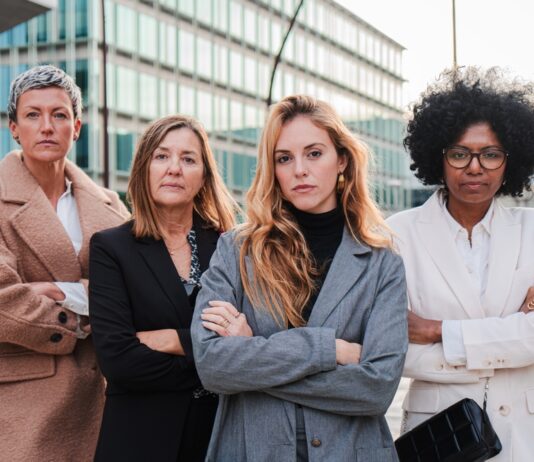 Gender Pay Gap: Darum sollten sich Frauen früh für ein gutes Gehalt stark machen Vier Geschäftsfrauen verschiedenen Alters und verschiedener Herkunft stehen mit verschränkten Armen nebeneinander und schauen entschlossen nach vorn.