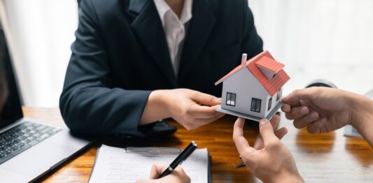 Maklervertrag für Kaufinteressierte: Was bedeutet das genau? Immobilienmakler überreicht ein Modell eines Einfamilienhauses, parallel wird ein Vertrag unterschrieben