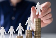 Mindestlohn: 10 Fragen und Antworten menschliche Hand, die eine kleine menschliche Figur auf die Erhöhung von Stackmünzen auf Holzschreibtisch legt