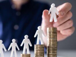 Mindestlohn: 10 Fragen und Antworten menschliche Hand, die eine kleine menschliche Figur auf die Erhöhung von Stackmünzen auf Holzschreibtisch legt