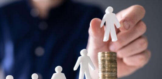 Mindestlohn: 10 Fragen und Antworten menschliche Hand, die eine kleine menschliche Figur auf die Erhöhung von Stackmünzen auf Holzschreibtisch legt