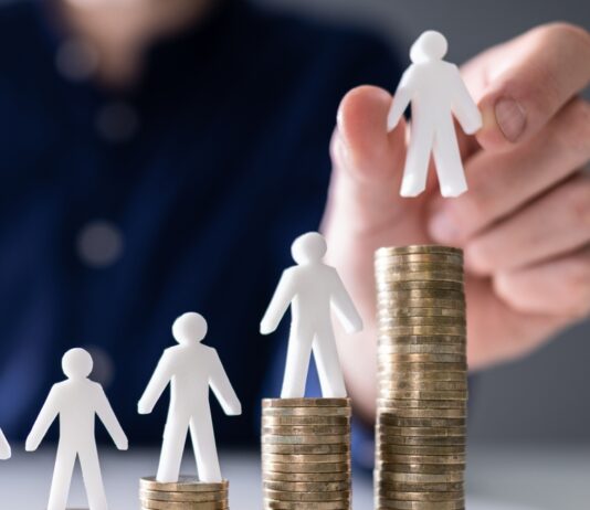 Mindestlohn: 10 Fragen und Antworten menschliche Hand, die eine kleine menschliche Figur auf die Erhöhung von Stackmünzen auf Holzschreibtisch legt