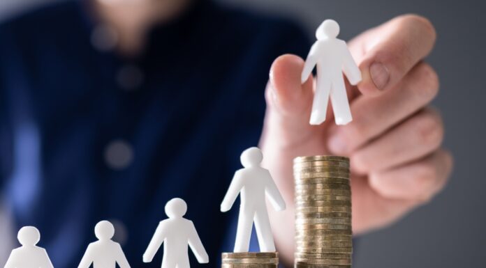 Mindestlohn: 10 Fragen und Antworten menschliche Hand, die eine kleine menschliche Figur auf die Erhöhung von Stackmünzen auf Holzschreibtisch legt