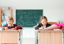 Von Schultüte bis Schnellhefter – was kostet die Einschulung? Ein Junge und ein Mädchen sitzen mit Schultüten auf den Tischen in einem Klassenraum. Im Hintergrund ist eine beschriebene Tafel zu sehen.