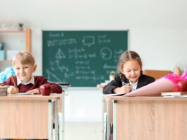 Von Schultüte bis Schnellhefter – was kostet die Einschulung? Ein Junge und ein Mädchen sitzen mit Schultüten auf den Tischen in einem Klassenraum. Im Hintergrund ist eine beschriebene Tafel zu sehen.