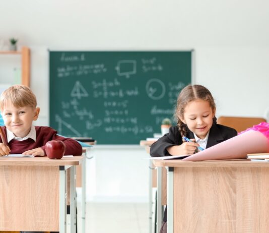 Von Schultüte bis Schnellhefter – was kostet die Einschulung? Ein Junge und ein Mädchen sitzen mit Schultüten auf den Tischen in einem Klassenraum. Im Hintergrund ist eine beschriebene Tafel zu sehen.