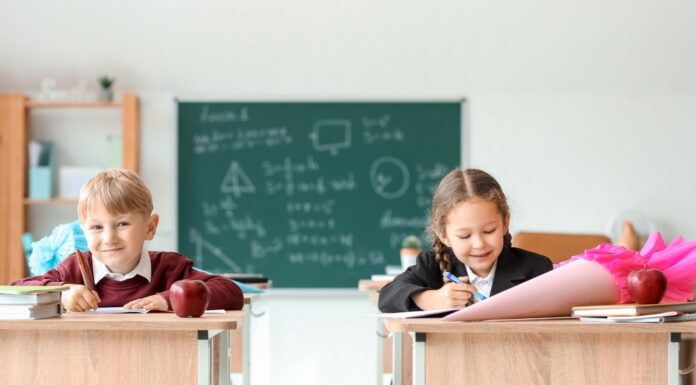 Von Schultüte bis Schnellhefter – was kostet die Einschulung? Ein Junge und ein Mädchen sitzen mit Schultüten auf den Tischen in einem Klassenraum. Im Hintergrund ist eine beschriebene Tafel zu sehen.
