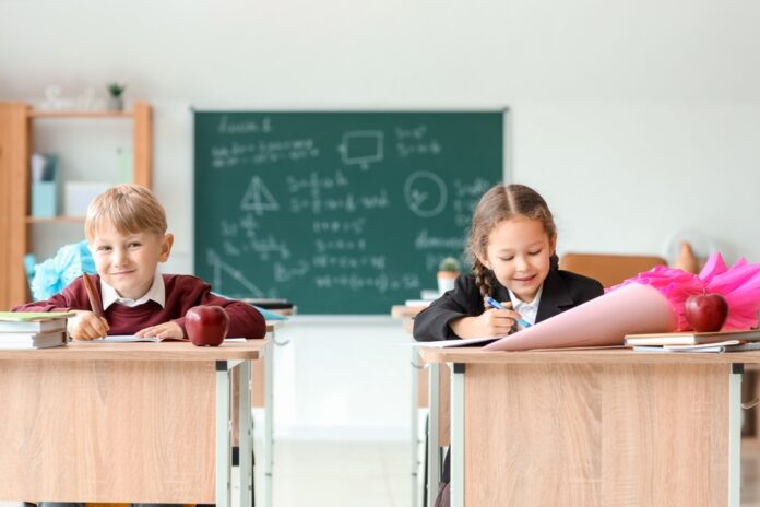 Ein Junge und ein Mädchen sitzen mit Schultüten auf den Tischen in einem Klassenraum. Im Hintergrund ist eine beschriebene Tafel zu sehen.