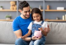 Vermögensbarometer 2025: Vertrauen wächst, Wissen bleibt Schlüssel Lächelndes kleines Mädchen steckt Münzen in ein rosa Sparschwein. Sie sitzt auf dem Schoß von ihrem Vater auf der Couch zu Hause.