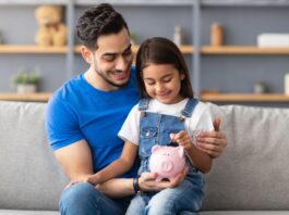 Vermögensbarometer 2025: Vertrauen wächst, Wissen bleibt Schlüssel Lächelndes kleines Mädchen steckt Münzen in ein rosa Sparschwein. Sie sitzt auf dem Schoß von ihrem Vater auf der Couch zu Hause.