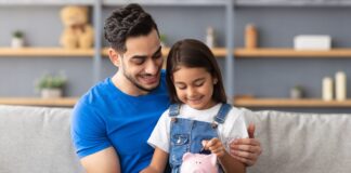 Vermögensbarometer 2025: Vertrauen wächst, Wissen bleibt Schlüssel Lächelndes kleines Mädchen steckt Münzen in ein rosa Sparschwein. Sie sitzt auf dem Schoß von ihrem Vater auf der Couch zu Hause.