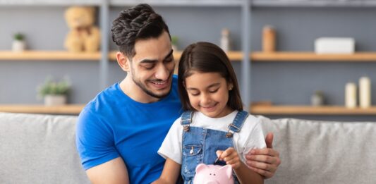 Vermögensbarometer 2025: Vertrauen wächst, Wissen bleibt Schlüssel Lächelndes kleines Mädchen steckt Münzen in ein rosa Sparschwein. Sie sitzt auf dem Schoß von ihrem Vater auf der Couch zu Hause.