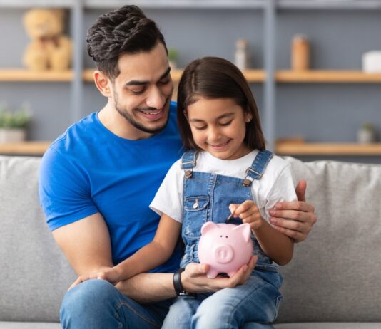 Vermögensbarometer 2025: Vertrauen wächst, Wissen bleibt Schlüssel Lächelndes kleines Mädchen steckt Münzen in ein rosa Sparschwein. Sie sitzt auf dem Schoß von ihrem Vater auf der Couch zu Hause.
