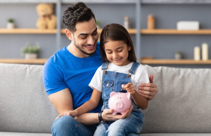 Lächelndes kleines Mädchen steckt Münzen in ein rosa Sparschwein. Sie sitzt auf dem Schoß von ihrem Vater auf der Couch zu Hause.