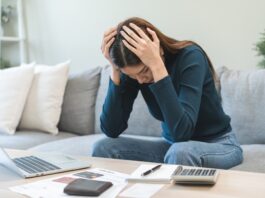 Schulden schnell abbauen: Tipps & Tricks Frau sitzt zuhause auf dem Sofa und stützt verzweifelt den Kopf in die Hände. Vor ihr auf dem Tisch stehen ein aufgeklapptes MacBook, Kreditkarte, Notizblock und Taschenrechner.