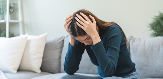 Schulden schnell abbauen: Tipps & Tricks Frau sitzt zuhause auf dem Sofa und stützt verzweifelt den Kopf in die Hände. Vor ihr auf dem Tisch stehen ein aufgeklapptes MacBook, Kreditkarte, Notizblock und Taschenrechner.