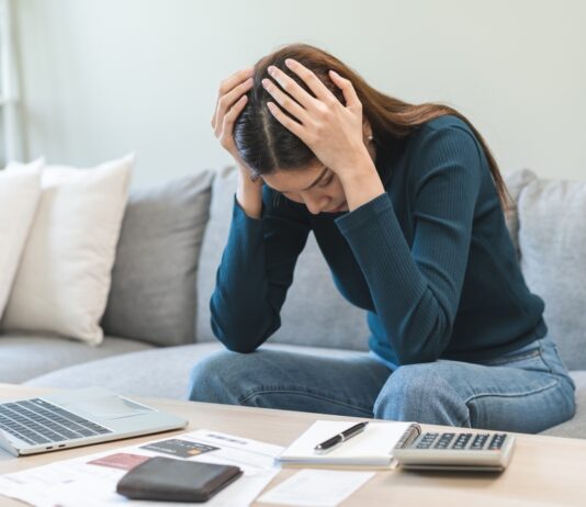 Schulden schnell abbauen: Tipps & Tricks Frau sitzt zuhause auf dem Sofa und stützt verzweifelt den Kopf in die Hände. Vor ihr auf dem Tisch stehen ein aufgeklapptes MacBook, Kreditkarte, Notizblock und Taschenrechner.