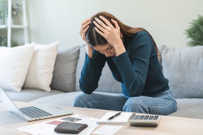 Frau sitzt zuhause auf dem Sofa und stützt verzweifelt den Kopf in die Hände. Vor ihr auf dem Tisch stehen ein aufgeklapptes MacBook, Kreditkarte, Notizblock und Taschenrechner.