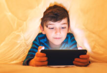 In-App- und In-Game-Käufe: Vorsicht Kostenfalle! Ein glücklich lächelnder Junge liegt bäuchlings unter einer Decke im Bett und spielt auf einem Smartphone in einem Spiel im Dunkeln. Das Gesicht des Kindes wird von einem hellen Monitor beleuchtet.