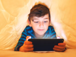 In-App- und In-Game-Käufe: Vorsicht Kostenfalle! Ein glücklich lächelnder Junge liegt bäuchlings unter einer Decke im Bett und spielt auf einem Smartphone in einem Spiel im Dunkeln. Das Gesicht des Kindes wird von einem hellen Monitor beleuchtet.