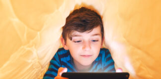 In-App- und In-Game-Käufe: Vorsicht Kostenfalle! Ein glücklich lächelnder Junge liegt bäuchlings unter einer Decke im Bett und spielt auf einem Smartphone in einem Spiel im Dunkeln. Das Gesicht des Kindes wird von einem hellen Monitor beleuchtet.