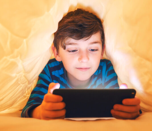 In-App- und In-Game-Käufe: Vorsicht Kostenfalle! Ein glücklich lächelnder Junge liegt bäuchlings unter einer Decke im Bett und spielt auf einem Smartphone in einem Spiel im Dunkeln. Das Gesicht des Kindes wird von einem hellen Monitor beleuchtet.