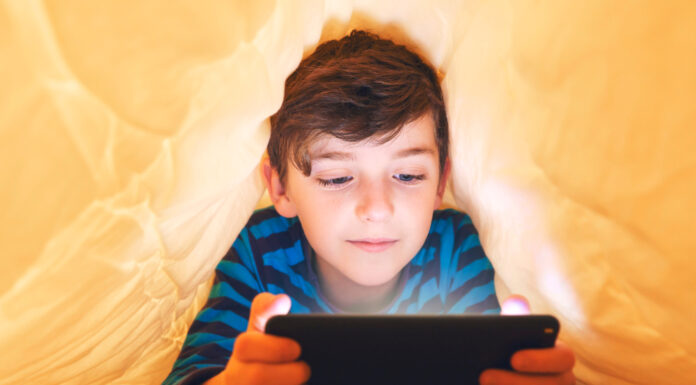 In-App- und In-Game-Käufe: Vorsicht Kostenfalle! Ein glücklich lächelnder Junge liegt bäuchlings unter einer Decke im Bett und spielt auf einem Smartphone in einem Spiel im Dunkeln. Das Gesicht des Kindes wird von einem hellen Monitor beleuchtet.