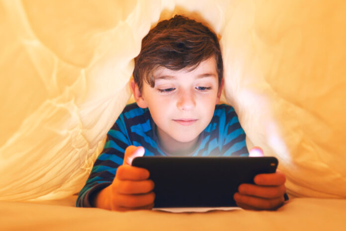 Ein glücklich lächelnder Junge liegt bäuchlings unter einer Decke im Bett und spielt auf einem Smartphone in einem Spiel im Dunkeln. Das Gesicht des Kindes wird von einem hellen Monitor beleuchtet.