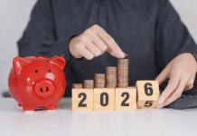 Das ändert sich 2026 Eine Person in einem dunkelgrauen Hemd legt eine Münze auf einen wachsenden Stapel aus Kupfermünzen, die auf Holzklötzen mit der Jahreszahl 2025 angeordnet sind. Daneben steht ein rotes Sparschwein auf einem Tisch, im Hintergrund liegt ein Taschenrechner – Symbol für Sparen und finanzielle Planung.