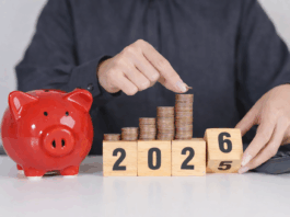 Das ändert sich 2026 Eine Person in einem dunkelgrauen Hemd legt eine Münze auf einen wachsenden Stapel aus Kupfermünzen, die auf Holzklötzen mit der Jahreszahl 2025 angeordnet sind. Daneben steht ein rotes Sparschwein auf einem Tisch, im Hintergrund liegt ein Taschenrechner – Symbol für Sparen und finanzielle Planung.