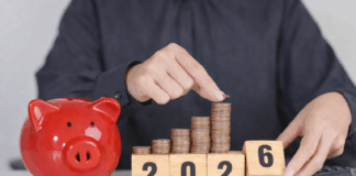 Das ändert sich 2026 Eine Person in einem dunkelgrauen Hemd legt eine Münze auf einen wachsenden Stapel aus Kupfermünzen, die auf Holzklötzen mit der Jahreszahl 2025 angeordnet sind. Daneben steht ein rotes Sparschwein auf einem Tisch, im Hintergrund liegt ein Taschenrechner – Symbol für Sparen und finanzielle Planung.