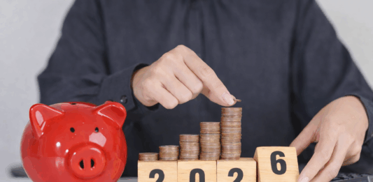 Das ändert sich 2026 Eine Person in einem dunkelgrauen Hemd legt eine Münze auf einen wachsenden Stapel aus Kupfermünzen, die auf Holzklötzen mit der Jahreszahl 2025 angeordnet sind. Daneben steht ein rotes Sparschwein auf einem Tisch, im Hintergrund liegt ein Taschenrechner – Symbol für Sparen und finanzielle Planung.