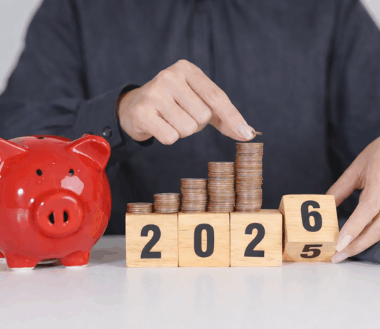 Das ändert sich 2026 Eine Person in einem dunkelgrauen Hemd legt eine Münze auf einen wachsenden Stapel aus Kupfermünzen, die auf Holzklötzen mit der Jahreszahl 2025 angeordnet sind. Daneben steht ein rotes Sparschwein auf einem Tisch, im Hintergrund liegt ein Taschenrechner – Symbol für Sparen und finanzielle Planung.