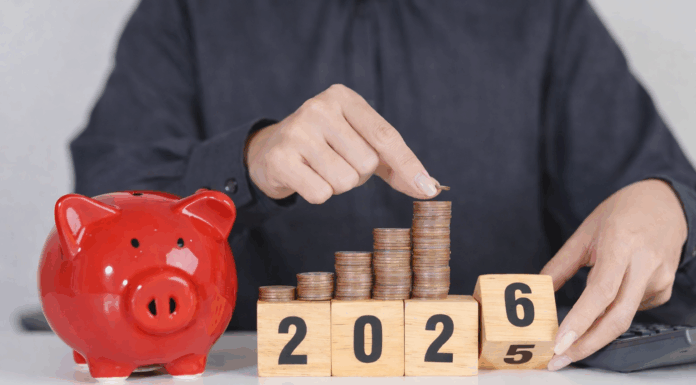 Das ändert sich 2026 Eine Person in einem dunkelgrauen Hemd legt eine Münze auf einen wachsenden Stapel aus Kupfermünzen, die auf Holzklötzen mit der Jahreszahl 2025 angeordnet sind. Daneben steht ein rotes Sparschwein auf einem Tisch, im Hintergrund liegt ein Taschenrechner – Symbol für Sparen und finanzielle Planung.