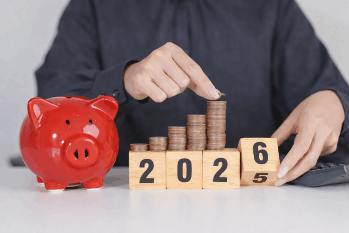 Eine Person in einem dunkelgrauen Hemd legt eine Münze auf einen wachsenden Stapel aus Kupfermünzen, die auf Holzklötzen mit der Jahreszahl 2025 angeordnet sind. Daneben steht ein rotes Sparschwein auf einem Tisch, im Hintergrund liegt ein Taschenrechner – Symbol für Sparen und finanzielle Planung.