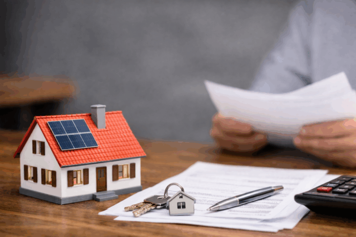 Modell eines Einfamilienhauses mit Solarpanel auf dem Dach, daneben Hausschlüssel, Vertragsunterlagen, Stift und Taschenrechner auf einem Holztisch.