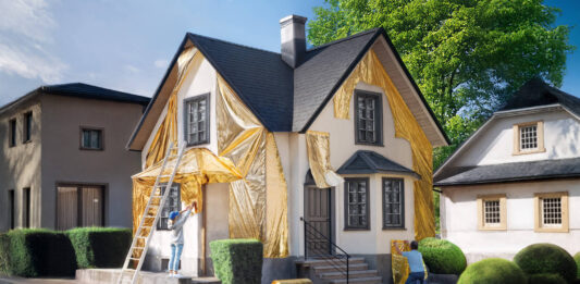 Modernisierungsrechner: Kosten berechnen und Förderungen nutzen Ein weißes Einfamilienhaus wird von einer Frau und einem Mann mit goldener Folie verkleidet. Vorn am Haus lehnt eine weiße Leiter.