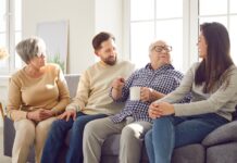 In jedem Alter: Was Sie Ihre Eltern über Geld fragen sollten Fröhlich lächelnde ältere Eltern und ihre erwachsenen Kinder Mann und Frau verbringen Zeit zusammen sitzen auf einem Sofa im Wohnzimmer zu Hause und sprechen mit einer Tasse Tee.