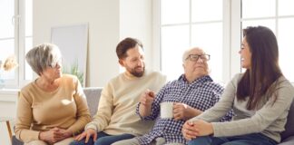 In jedem Alter: Was Sie Ihre Eltern über Geld fragen sollten Fröhlich lächelnde ältere Eltern und ihre erwachsenen Kinder Mann und Frau verbringen Zeit zusammen sitzen auf einem Sofa im Wohnzimmer zu Hause und sprechen mit einer Tasse Tee.