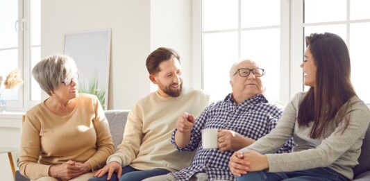 In jedem Alter: Was Sie Ihre Eltern über Geld fragen sollten Fröhlich lächelnde ältere Eltern und ihre erwachsenen Kinder Mann und Frau verbringen Zeit zusammen sitzen auf einem Sofa im Wohnzimmer zu Hause und sprechen mit einer Tasse Tee.
