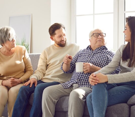 In jedem Alter: Was Sie Ihre Eltern über Geld fragen sollten Fröhlich lächelnde ältere Eltern und ihre erwachsenen Kinder Mann und Frau verbringen Zeit zusammen sitzen auf einem Sofa im Wohnzimmer zu Hause und sprechen mit einer Tasse Tee.