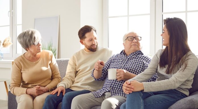 In jedem Alter: Was Sie Ihre Eltern über Geld fragen sollten Fröhlich lächelnde ältere Eltern und ihre erwachsenen Kinder Mann und Frau verbringen Zeit zusammen sitzen auf einem Sofa im Wohnzimmer zu Hause und sprechen mit einer Tasse Tee.