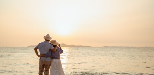 Rente im Ausland: Was Sie alles wissen sollten Ein Paar, das bei Sonnenuntergang am Strand steht, wobei der Mann die Hand der Frau hält.