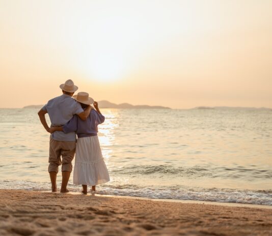 Rente im Ausland: Was Sie alles wissen sollten Ein Paar, das bei Sonnenuntergang am Strand steht, wobei der Mann die Hand der Frau hält.
