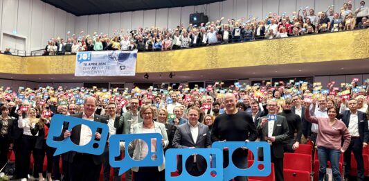 Wir sagen „JO!“ zu den Olympischen Spielen in Kiel Viele Menschen stehen im Konzertsaal des Kieler Schlosses und halten bunte Schilder hoch, auf denen "JO!" steht.