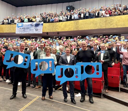 Wir sagen „JO!“ zu den Olympischen Spielen in Kiel Viele Menschen stehen im Konzertsaal des Kieler Schlosses und halten bunte Schilder hoch, auf denen "JO!" steht.