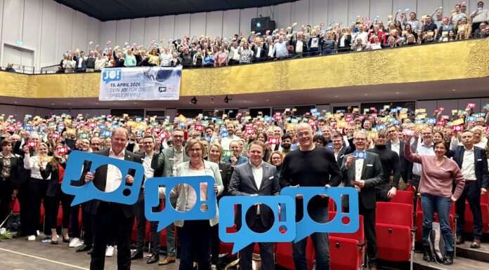Wir sagen „JO!“ zu den Olympischen Spielen in Kiel Viele Menschen stehen im Konzertsaal des Kieler Schlosses und halten bunte Schilder hoch, auf denen "JO!" steht.