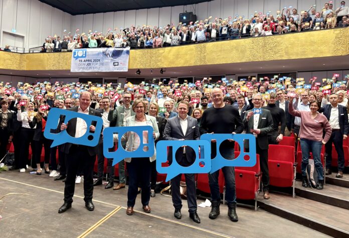 Viele Menschen stehen im Konzertsaal des Kieler Schlosses und halten bunte Schilder hoch, auf denen "JO!" steht.