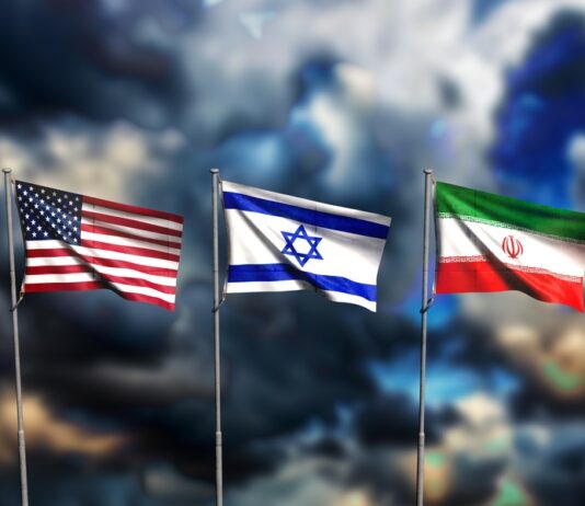 Iran-Krieg: Das wird nun teurer Die Flaggen von den USA, Isreal und Iran wehen vor einem dunklen, wolkenverhangenen Himmel.